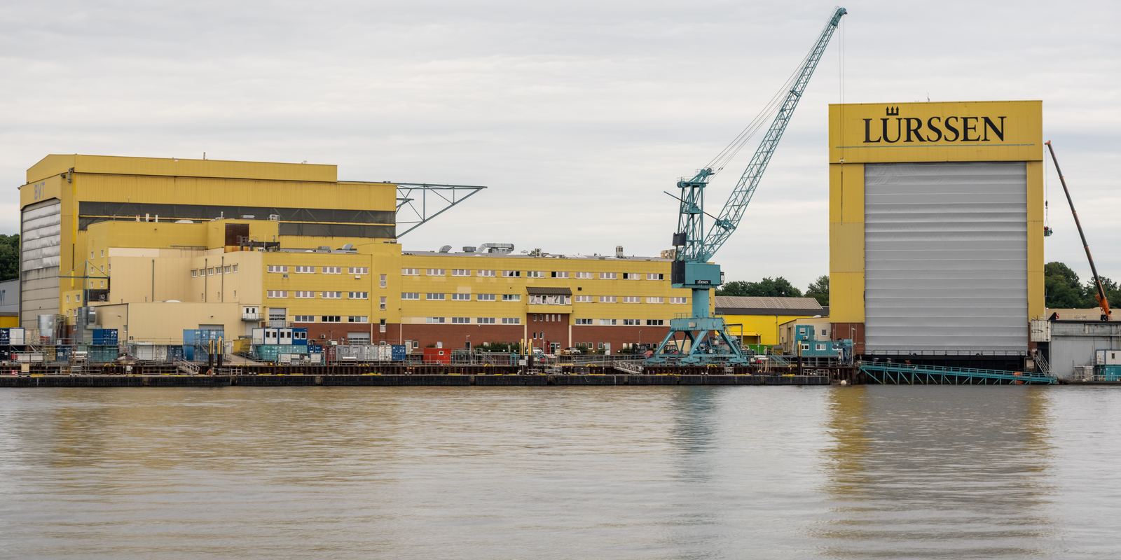 Large yellow construction halls and a crane at the Lürssen shipyard in Bremen-Vegesack beside the river