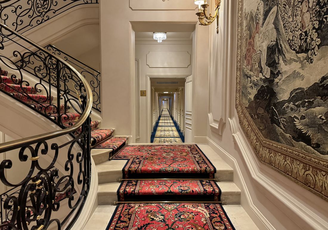 A curving staircase with a wrought iron rail and red patterned carpet inside the Ritz Paris